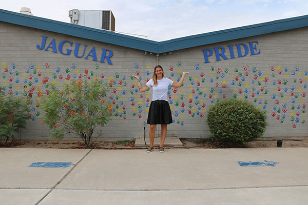 a woman stands with her arms up; behind her, a wall covered in colorful handprints reads "Jaguar Pride"
