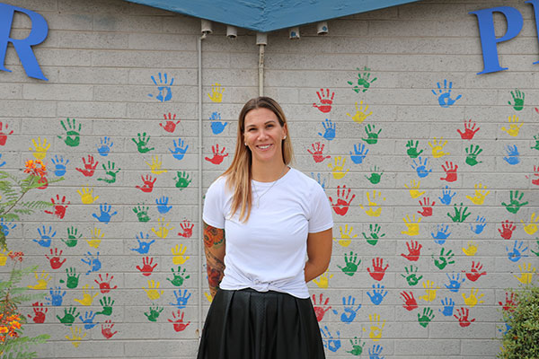 a woman smiles as she stands with her back to a wall covered in colorful handprints
