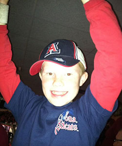A young boy, wearing a baseball cap and jersey, raises his hands in the air