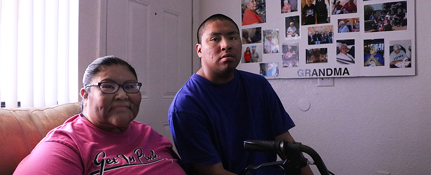 A mom sits on a couch with a walker in front of her. Her son sits beside her on the arm of the couch.