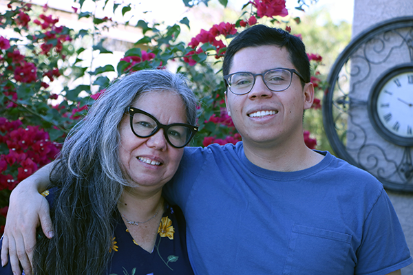 Mother and son, with his arm around her, stand outside the entrance to their house