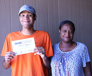 a young man wearing a baseball cap holds a certificate in front of him; an older woman stands beside him, smiling