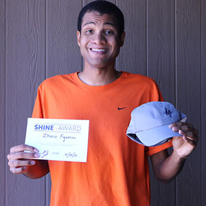 a young man smiles, holding a certificate in one hand and a baseball cap in the other