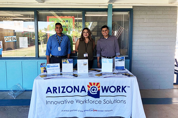 two men and a woman stand behind a cloth-covered booth that reads "ARIZONA@WORK Innovative Workforce Solutions"