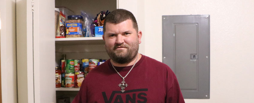 Close-shaven bearded man wearing a T-shirt stands in front of his kitchen pantry.