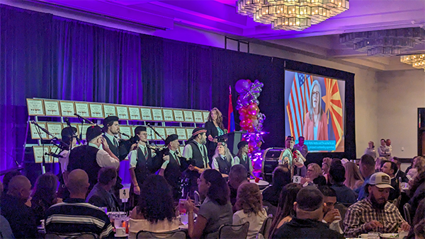 A group performing on stage at an event, with a screen displaying a person by flags and tables of people watching.