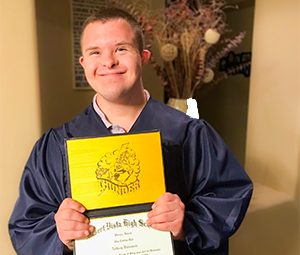 A young person in a blue graduation gown holding a yellow diploma cover with a lion crest.