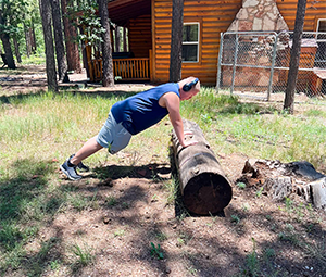 A person doing an inclined push-up on a log in a forest near a cabin.