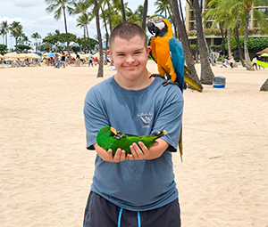 A young person holds a green parrot and has a blue and yellow macaw on their shoulder at a sandy beach with palm trees.