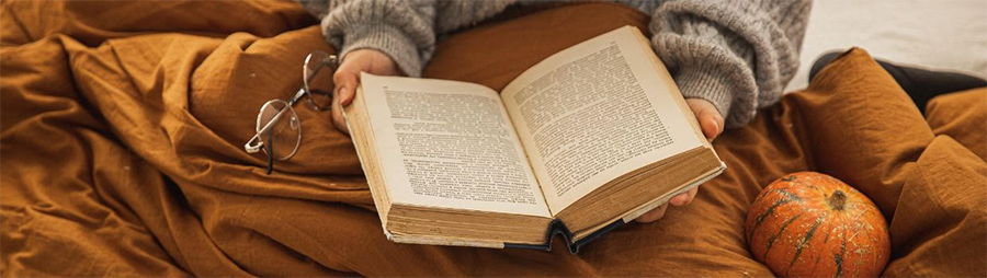 Person reading an open book on a rust-colored blanket with glasses and a pumpkin nearby.