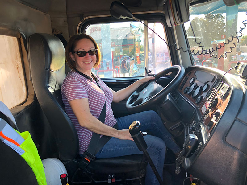 a woman sitting in the driver's seat of a large vehicle is smiling; a large steering wheel and many controls are visible