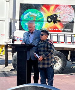 Man at podium with his arm around the shoulders of a teenage boy, gives a speech about the teen’s winning artwork