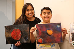 Mom and teen son stand side-by-side, each holding up one of the teen’s paintings