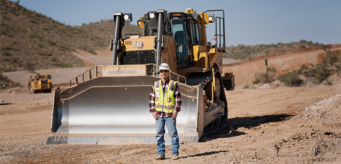 A person stands in front of a large yellow bulldozer on a dirt construction site with hills in the background.