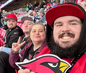 Four people in a stadium wearing sports apparel, one holding a cardinal foam cutout.