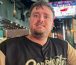 Person wearing a black and tan "Serpientes" baseball jersey, sitting indoors with a sports game on TV in the background.