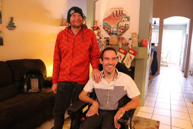 Man standing with his hand on the shoulder of his friend who is seated in a power wheel chair
