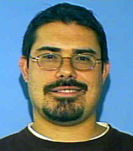 A white with man short, black hair, brown eyes, and a black goatee is wearing glasses and a black t-shirt.