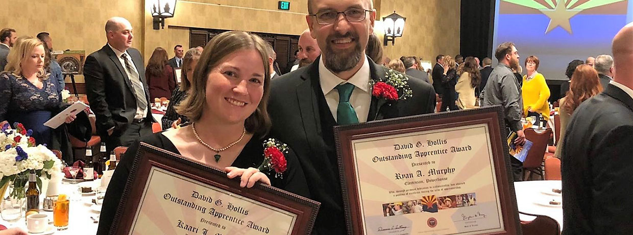 A man and woman stand side by side, smiling, each holding a framed certificate