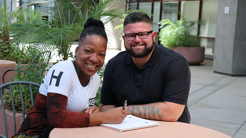 a woman and man smile at the camera while sitting at a table amongst potted plants