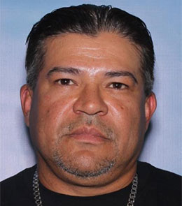 Man with brown eyes, slicked, black hair, and a white goatee is wearing a silver chain and black t-shirt