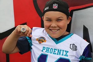 Boy wearing football jersey and ball cap holds up the MVP ring he was awarded.