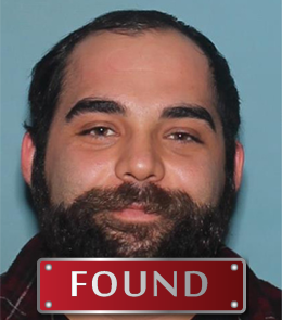 A man with brown eyes, black hair, and a beard is wearing a red, plaid, flannel shirt