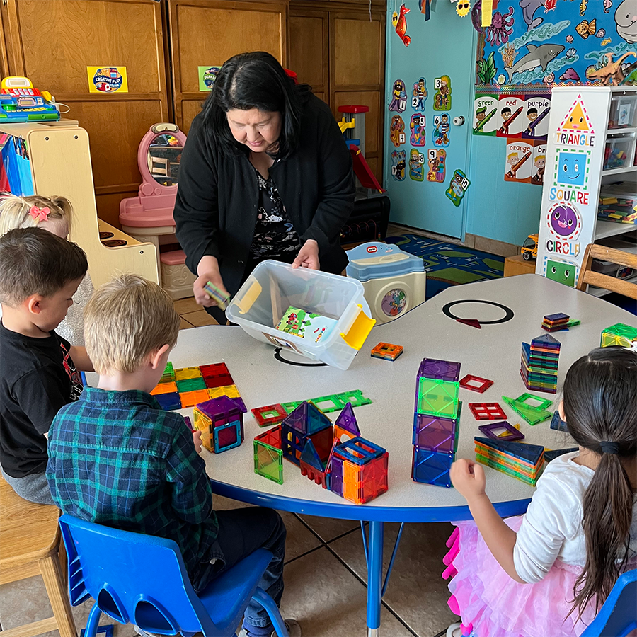 A woman and four children are building structures with colorful magnetic tiles at a round table in a classroom.