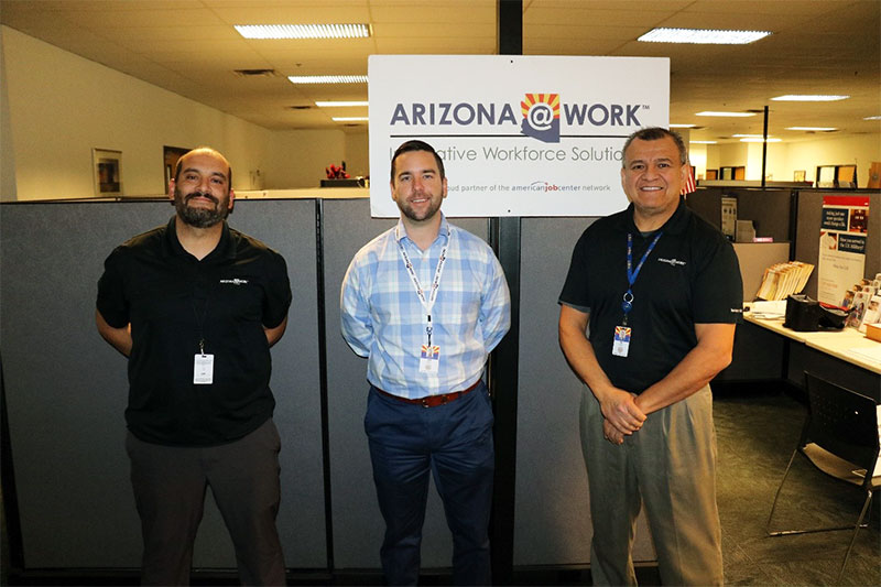 three men stand inside an office; in the background, an ARIZONA@WORK sign and cubicles are visible