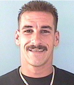 A white man with brown, spiked hair, brown eyes, and a mustache is wearing a gold chain and black t-shirt