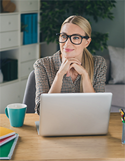 A person with glasses and long blonde hair sits at a desk with a laptop, a mug, notebooks, and pencils.