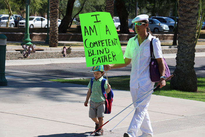 A blind man holds a sign that says "I am a competent blind father" as he walks with his young son  