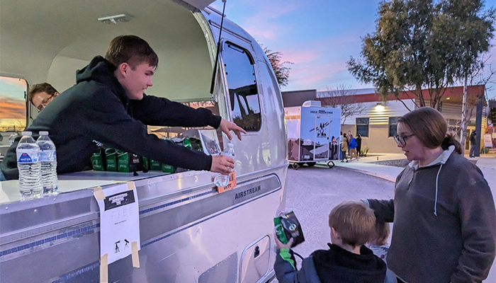 Person handing a bottled water from a silver Airstream trailer to a child and woman at twilight.