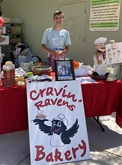 A person stands behind a red-covered table displaying baked goods and a sign for "Cravin' Ravens Bakery."