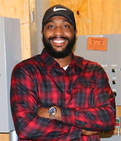 Person in a red and black checkered shirt and black cap, smiling with arms crossed in front of electrical panels.