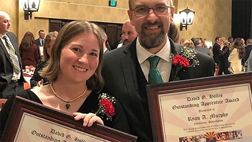 Two people in formal attire holding framed awards at a formal event.