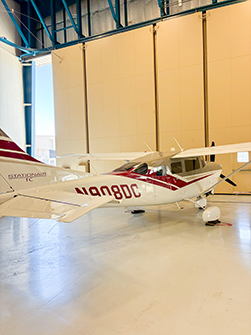 Small white airplane with maroon stripes parked inside a hangar.