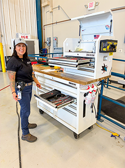 A woman in a workshop stands next to a large white equipment cart with tools and fire extinguishers.