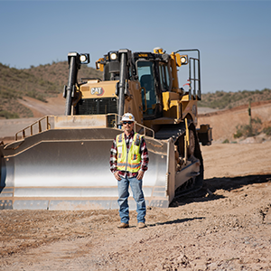 A person stands in front of a large yellow bulldozer on a dirt construction site with hills in the background.