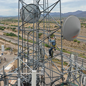 A worker climbs a telecommunications tower in a desert landscape with highways and distant mountains.