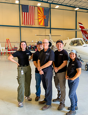 Five people standing in a hangar with an aircraft next to them. Five people standing in a hangar with an aircraft next to them.