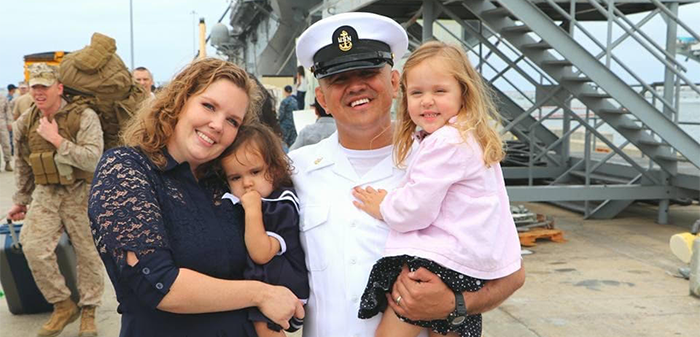 A naval officer in uniform with family on a dock, ship in background.