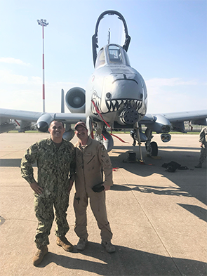 Two individuals in military uniforms stand in front of an A-10 Thunderbolt II with shark mouth nose art on an airstrip.