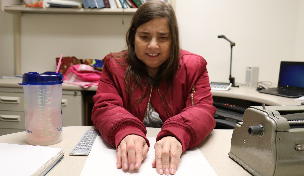 A woman in a red jacket reads a paper at a desk with various items, including a water bottle and braille notetaker.