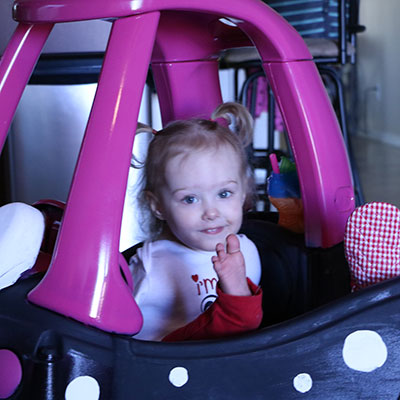 A toddler sitting in a toy car shaped like a lady bug, raises her right foot as a way to wave hello.