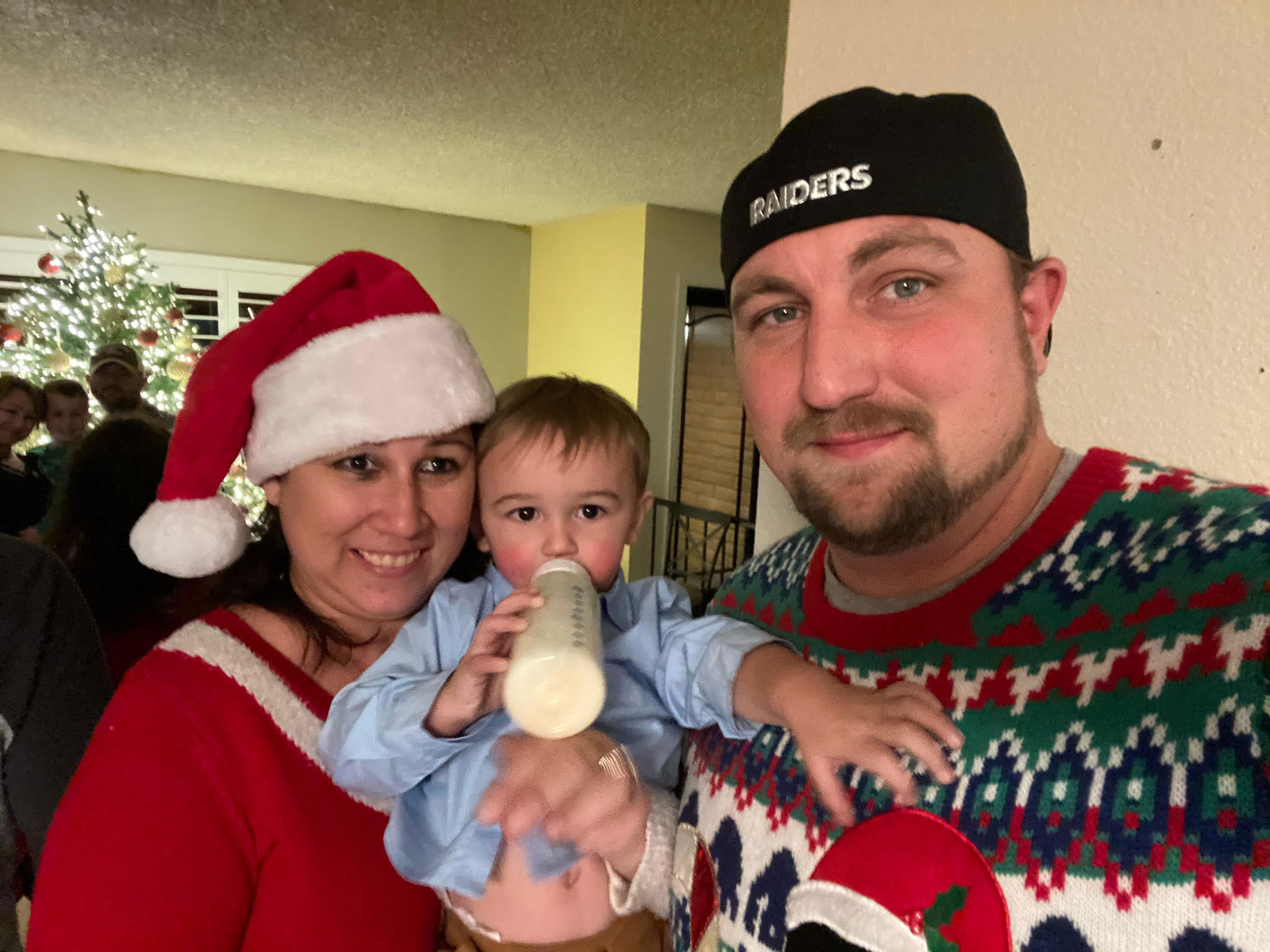 Dad, wearing a Raiders hat and Christmas sweater, Mom, wearing a Santa cap and red top, and their toddler son drinking from a bottle pose for a holiday photo