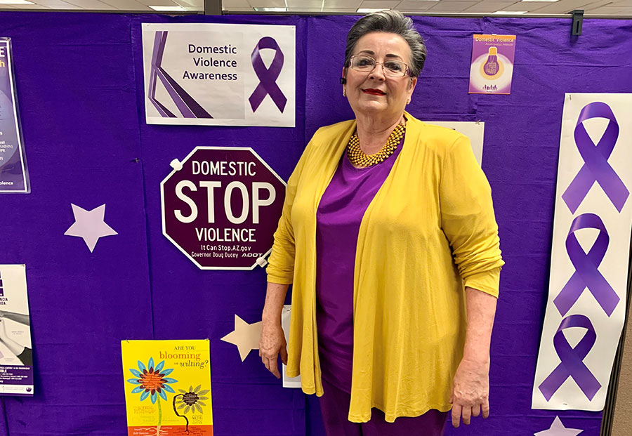 a woman dressed in yellow and purple poses for a photo; behind her, Domestic Violence awareness paraphernalia display on a purple wall