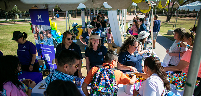 People gathered at an outdoor event under canopy tents with promotional materials on display. People gathered at an outdoor event under canopy tents with promotional materials on display.