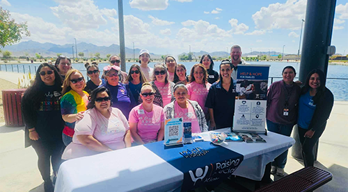 Group of people smiling at an outdoor event with a table displaying "Raising Special Kids" materials, under a sunny sky with clouds. Group of people smiling at an outdoor event with a table displaying "Raising Special Kids" materials, under a sunny sky with clouds.