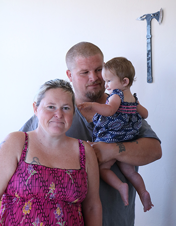 Mom, dad and daughter face the camera. Dad holds his 10-month-old girl who is having fun pulling her mother’s hair.